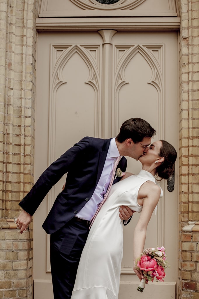 Hochzeitfotograf Potsdam Bräutigam küsst Braut innig vor beiger Backsteinmauer der Kirche