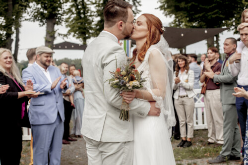 Hochzeitsfotograf Berlin Braut und Bräutigam üssen sich und Gäste applaudieren im Hintergrund in Richtershorn am See